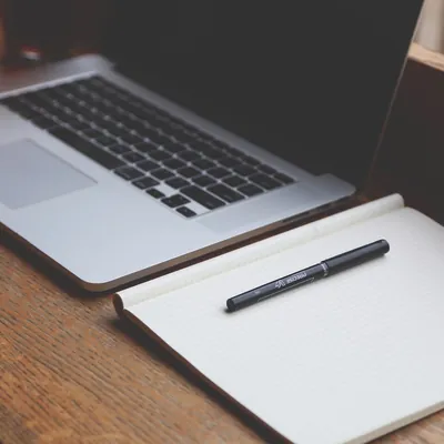 Laptop on a wooden desk beside an open grid notepad and pen