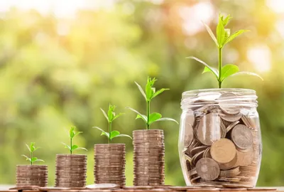 Stacks of coins growing taller from left to right, each topped with a seedling, beside a jar of coins